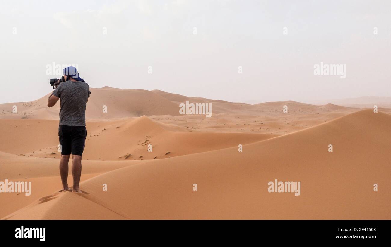 Un giovane in pantaloncini, che scatta foto da una grande duna di sabbia nel deserto del Sahara in Marocco. Foto Stock