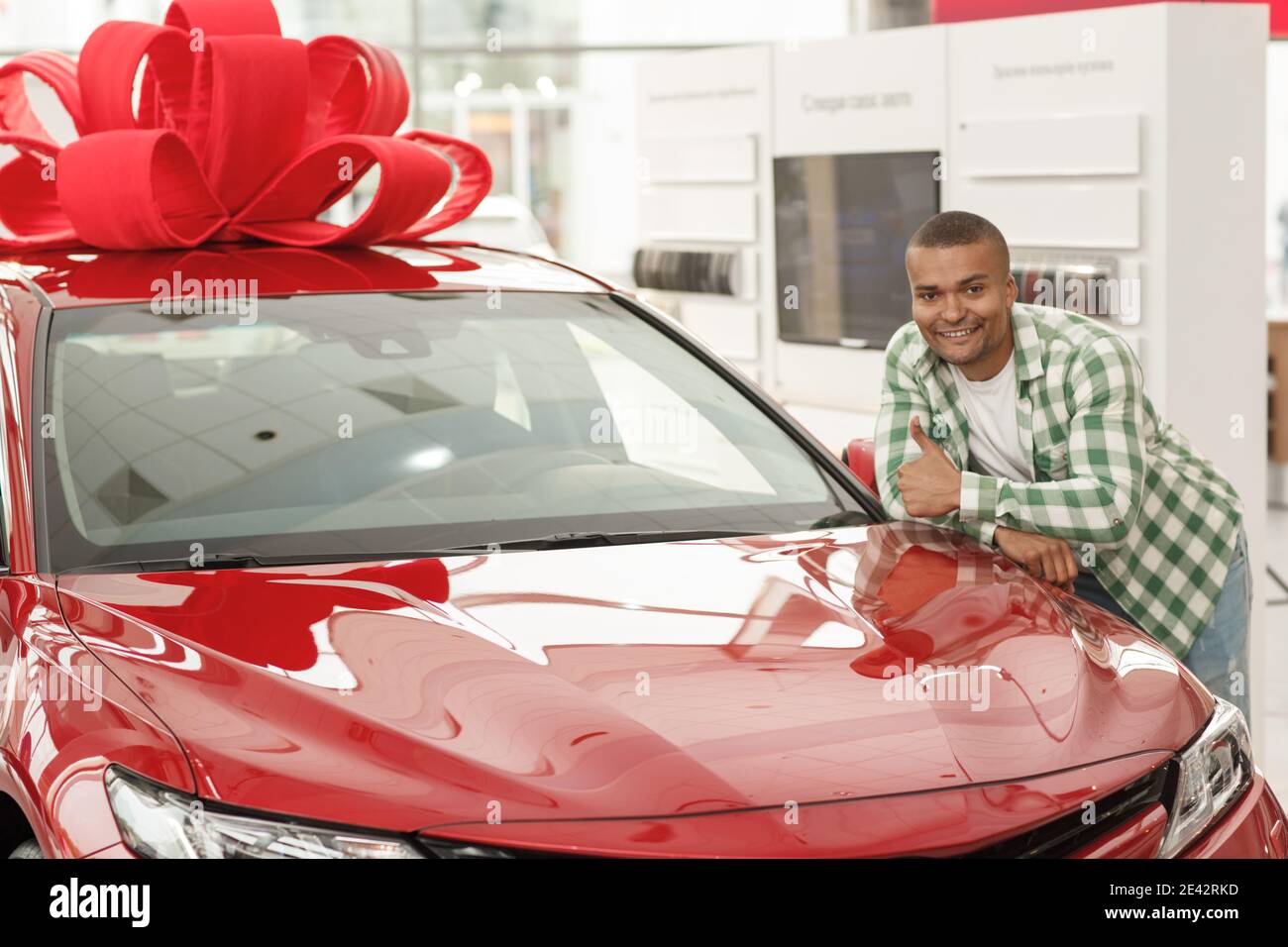 Ritratto orizzontale di un felice bel giovane africano sorridente alla macchina fotografica, mostrando i pollici in su, in posa vicino l'automobile appena comprato ai commercianti Foto Stock