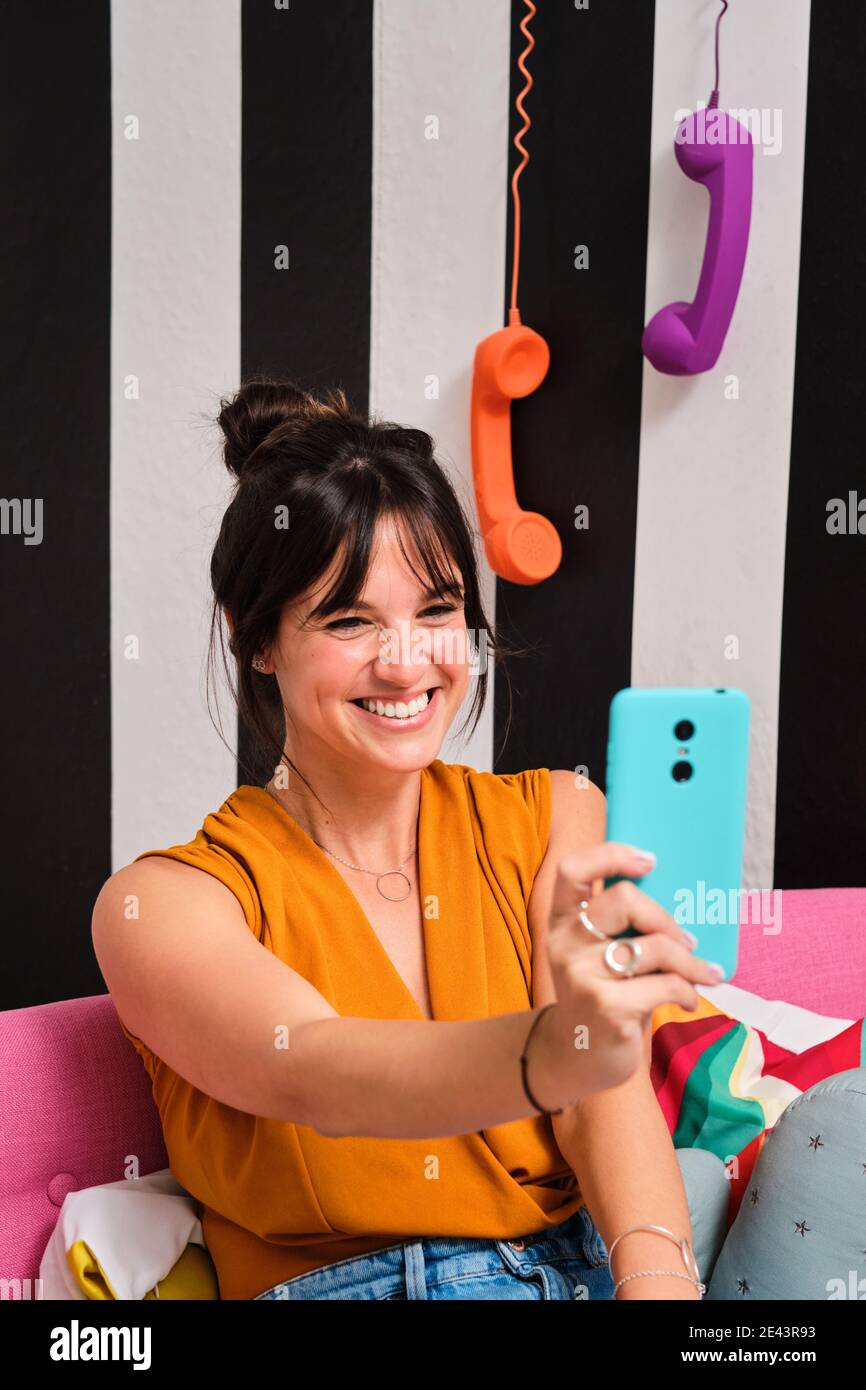 Allegra giovane femmina che si autoritratto su telefono cellulare blu mentre si siede sul divano in una camera elegante con pareti a righe e ricevitori telefonici retrò Foto Stock