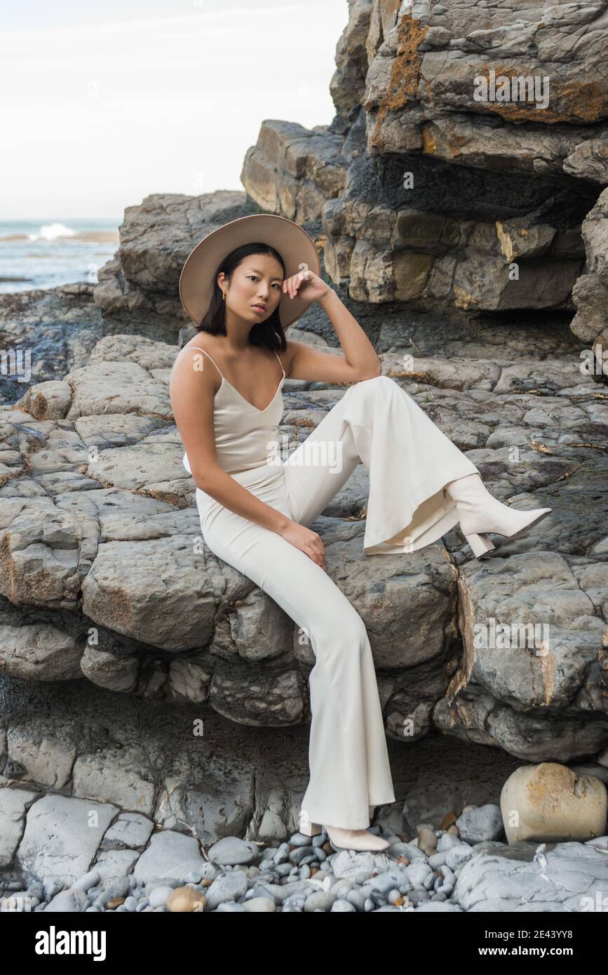 Corpo pieno di elegante giovane e sottile donna asiatica in bianco vestito e cappello guardando la macchina fotografica mentre si riposa sulla roccia costa vicino al mare Foto Stock