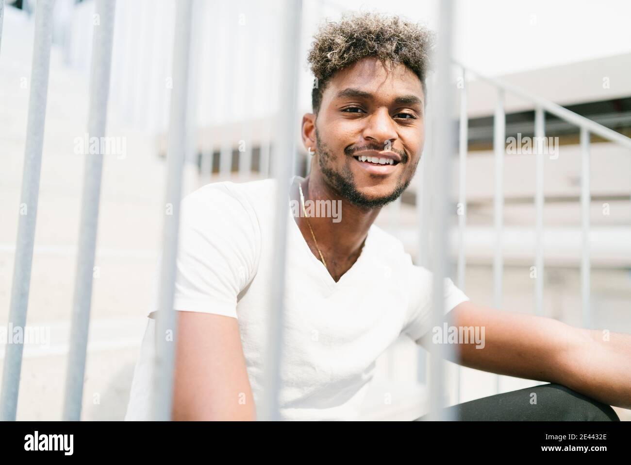 Bel ragazzo nero hipster con i capelli ricci vestito in camicia bianca casual guardando la macchina fotografica e sorridendo mentre seduto sulle scale vicino meta Foto Stock