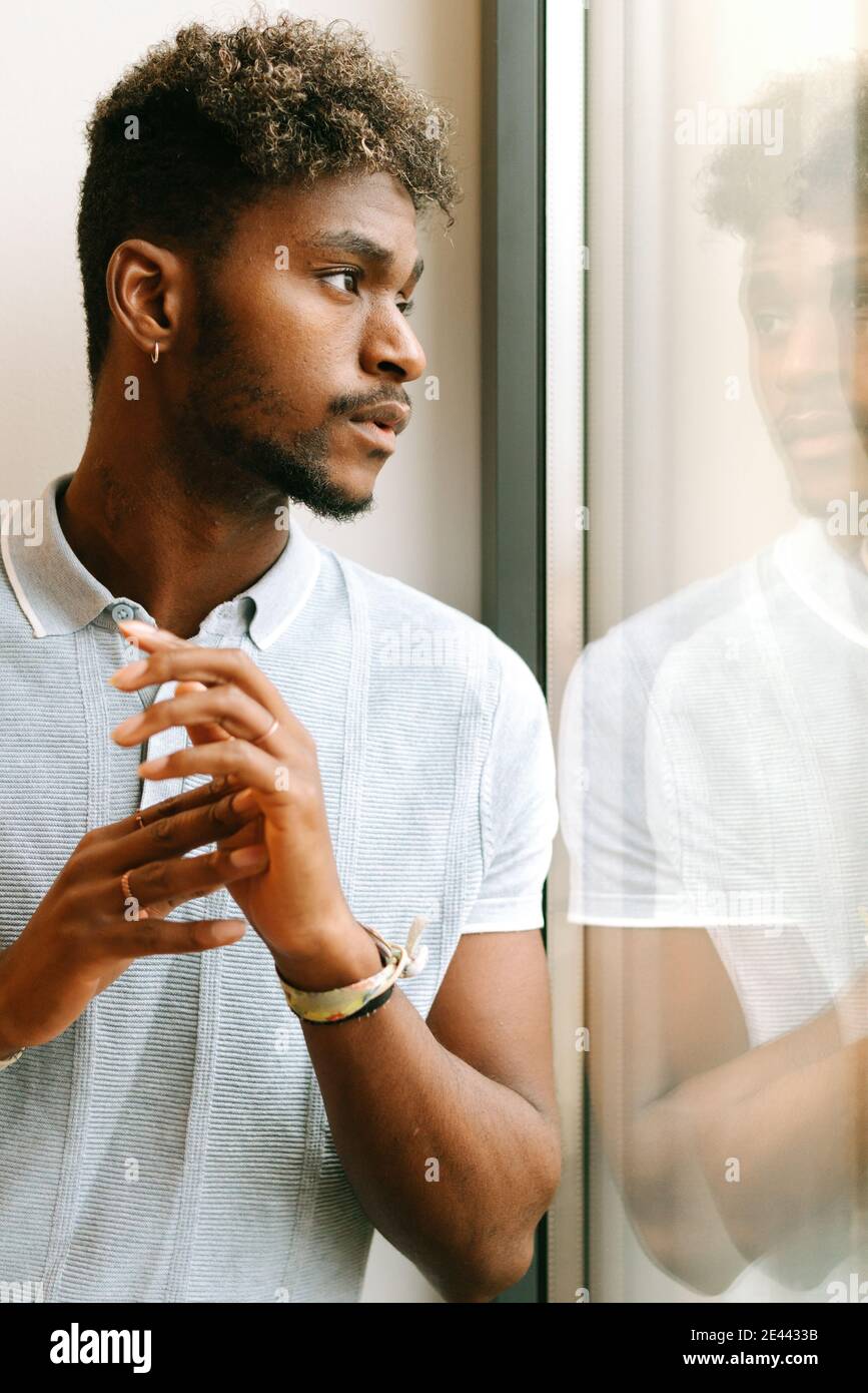 Serio giovane nero sopportato maschio con capelli ricci in casual camicia che guarda lontano pensieroso mentre si è in piedi vicino alla finestra Foto Stock