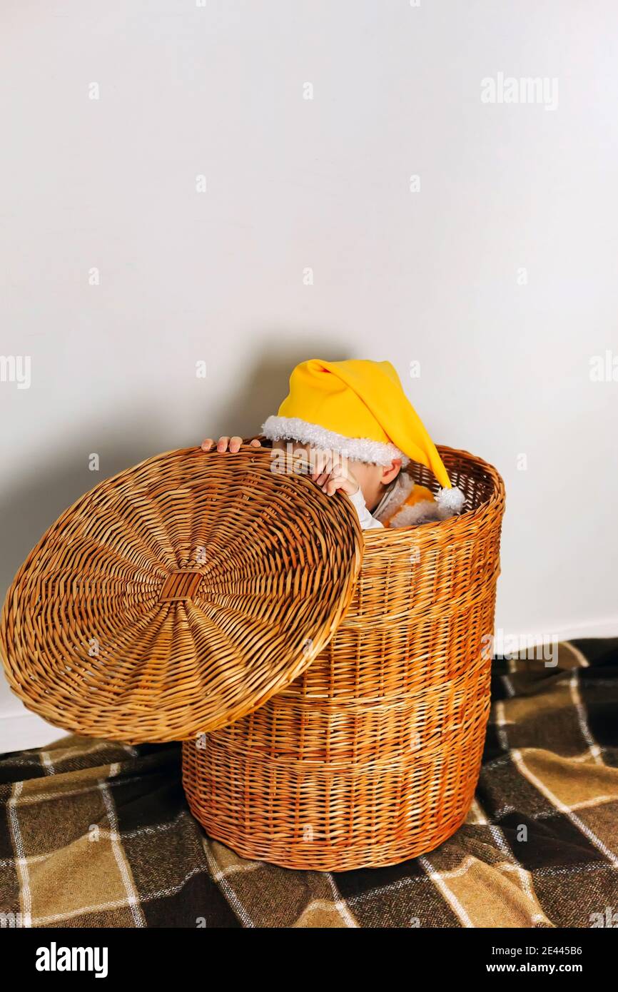 Cute baby boy sedersi in cestino di paglia e nascondere la faccia sotto il cappuccio, indossare giallo divertente elfo ghome cappello, famiglia divertimento a casa Foto Stock