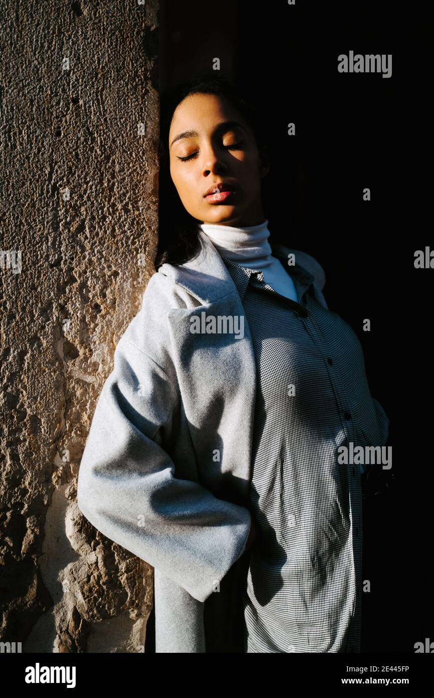 Serena giovane donna etnica in elegante camice grigio appoggiato alla schiena contro muro di pietra invecchiato e godendo della luce del sole Foto Stock