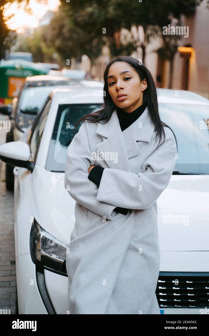 Elegante donna con maniche pieghevoli mentre si appoggia al moderno auto nel centro della città guardando la fotocamera Foto Stock