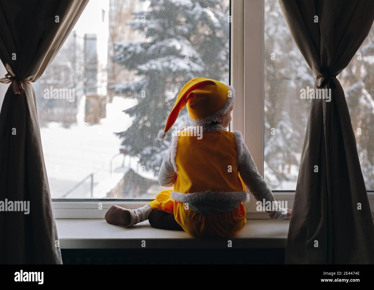 Bambino sedersi indietro nella finestra in elfo gnome vestito giallo e guardare la neve inverno che cade vista, felice tempo di famiglia a casa Foto Stock