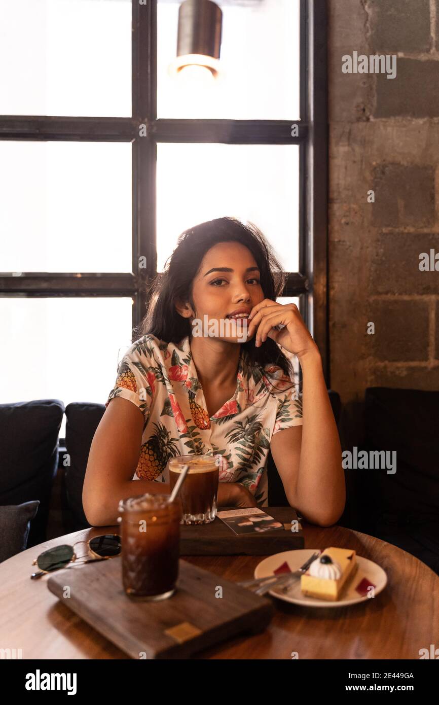 Felice giovane etnia indiana seduta a tavola con dolce dessert e bevande e guardando la macchina fotografica mentre si riposa caffè contro la finestra Foto Stock