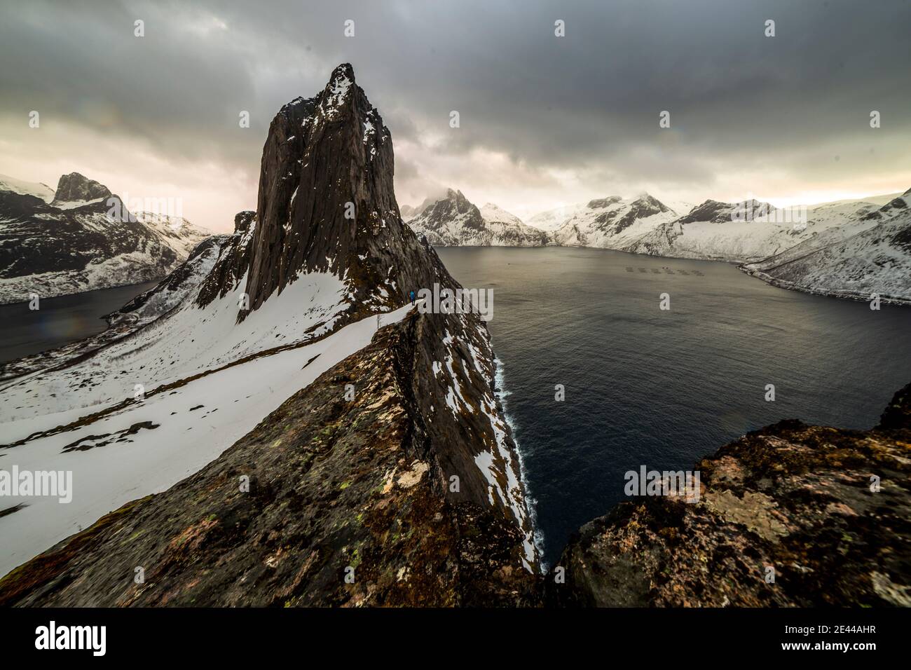 Paesaggio remoto del viaggiatore che cammina lungo il bordo roccioso della montagna con neve situata vicino al mare durante le escursioni in inverno in Norvegia Foto Stock