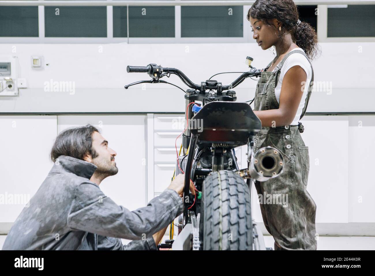 Diversi meccanici maschili e femminili nell'abbigliamento da lavoro che fissano la motocicletta insieme in garage luminoso Foto Stock