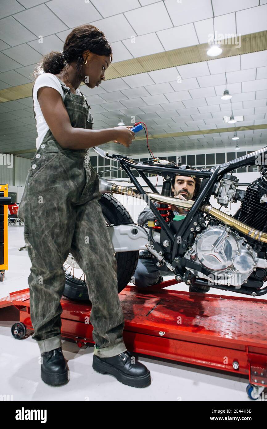 Diversi meccanici maschili e femminili nell'abbigliamento da lavoro che fissano la motocicletta insieme in garage luminoso Foto Stock