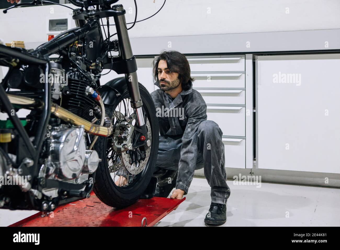 Serio tecnico maschile in abbigliamento da lavoro che si prepara per il fissaggio di moto moderne collocato su un ascensore in metallo in un'officina luminosa Foto Stock