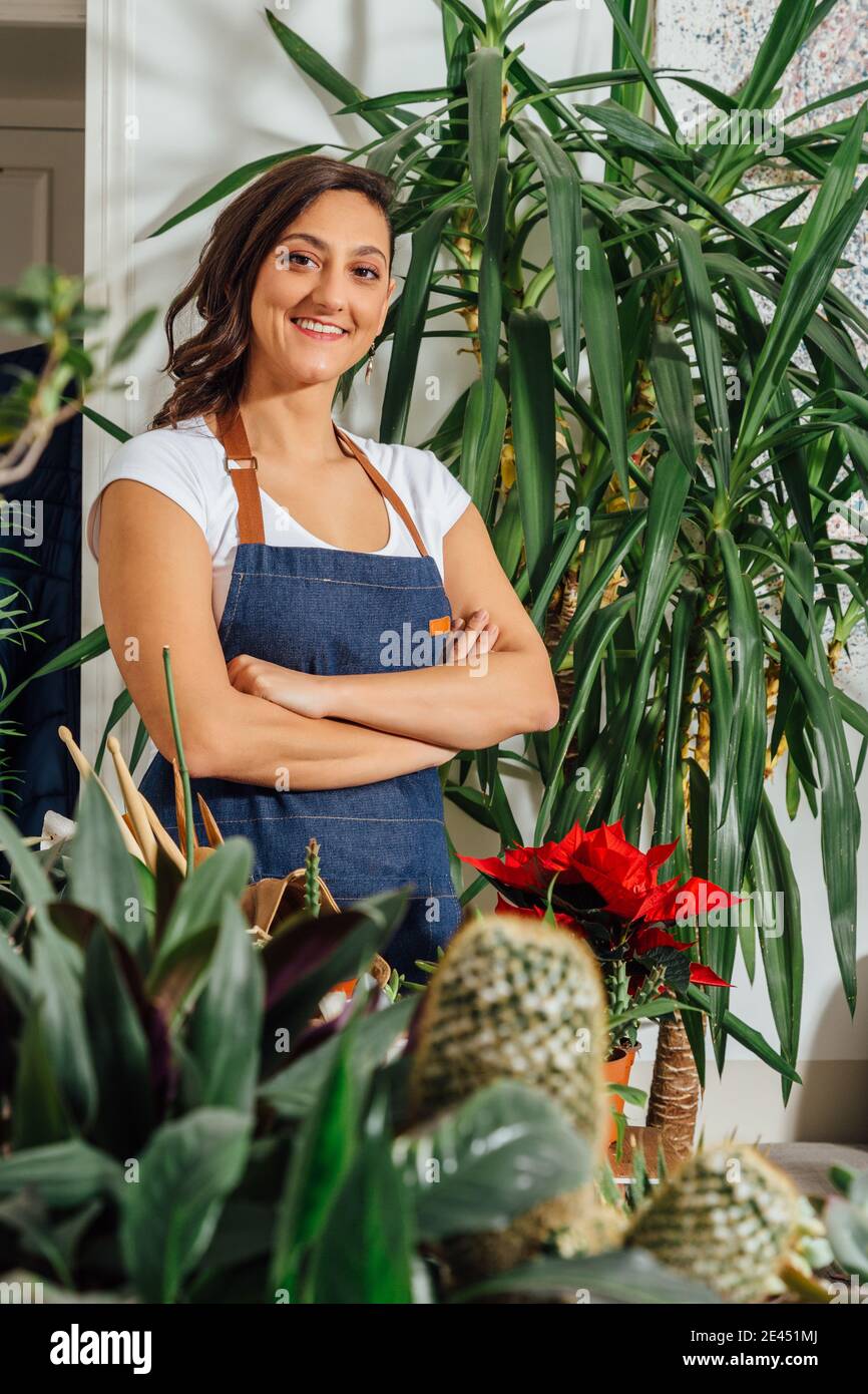 Sorridente giardiniere femminile in grembiule in piedi con braccia incrociate dentro negozio di fiori con piante in vaso assortite e guardando la macchina fotografica Foto Stock