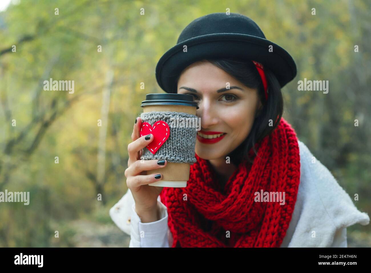 Giovane donna in un cappello nero beve caffè da una tazza accogliente sulla natura. Sorriso luminoso e buon umore. Foto Stock