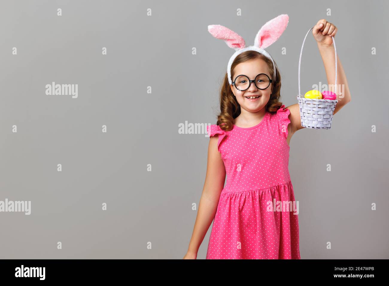 Felice ragazza carina che indossa orecchie coniglietto pasquale e occhiali su sfondo grigio. Il bambino sta tenendo un cestino di uova. Foto Stock