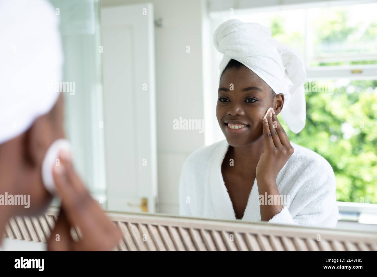 Donna afroamericana in accappatoio che rimuove il trucco mentre si guarda in lo specchio del bagno Foto Stock