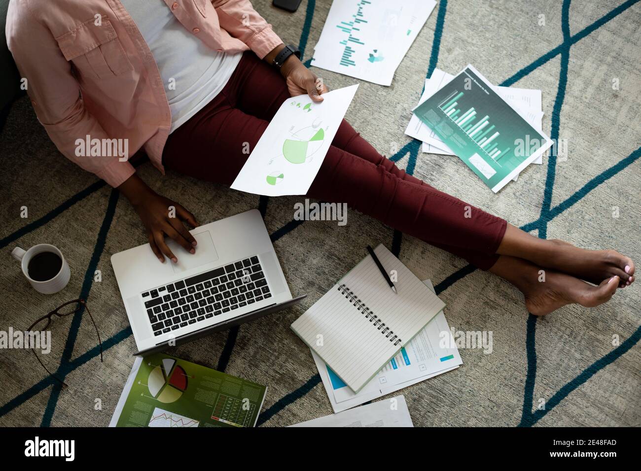 Donna afroamericana seduta sul pavimento usando il laptop che passa attraverso documenti da casa Foto Stock