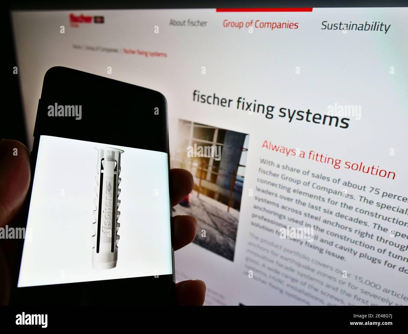 Persona che tiene uno smartphone con presa a muro sul display di fronte al sito del produttore Fischer Group (Fischerwerke). Mettere a fuoco lo schermo del telefono cellulare. Foto Stock