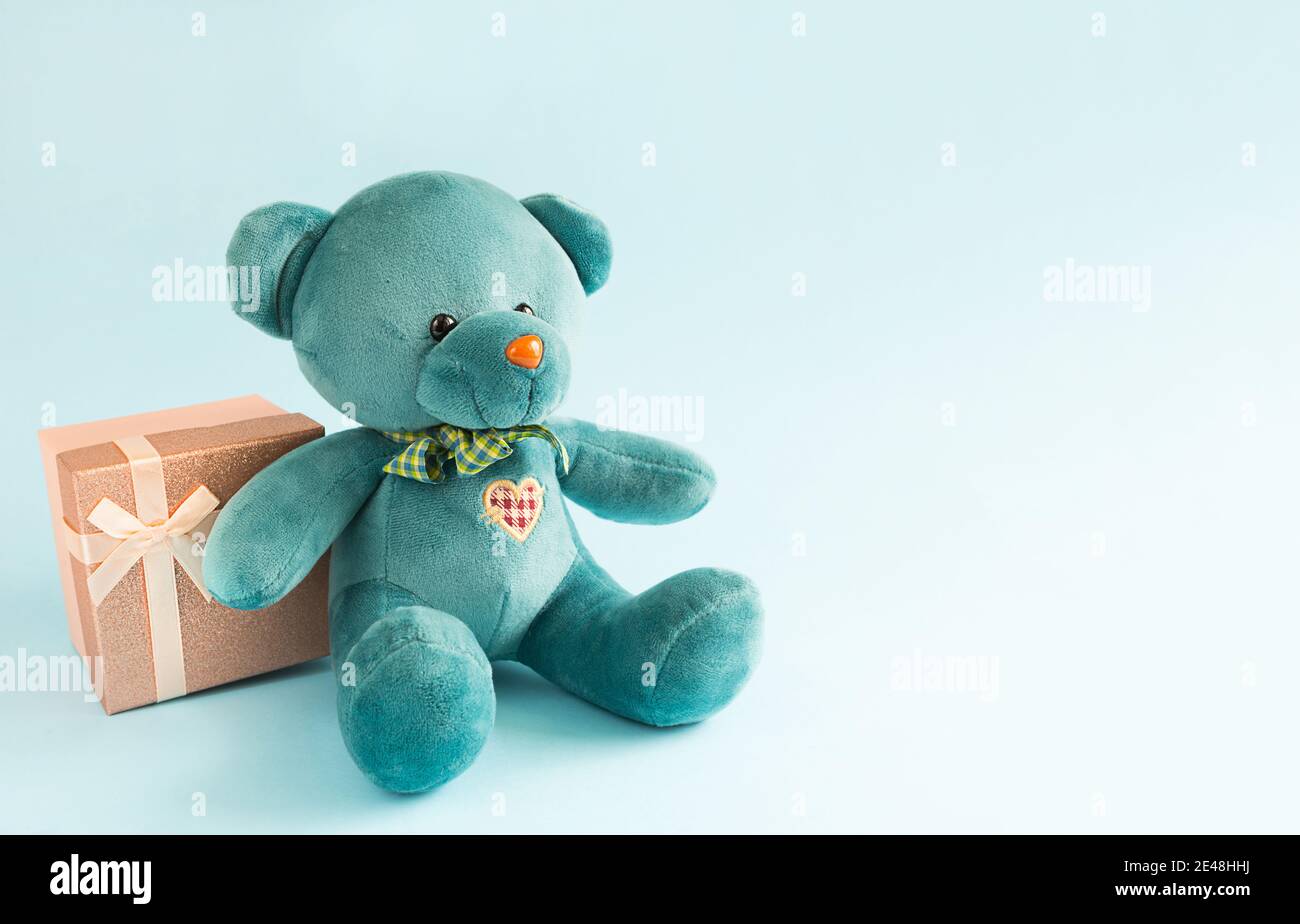 Un morbido orsacchiotto turchese con cuore ricamato contiene una scatola regalo e un prua su sfondo blu. Giocattolo per bambini. Amore, un regalo per le vacanze, un declar Foto Stock