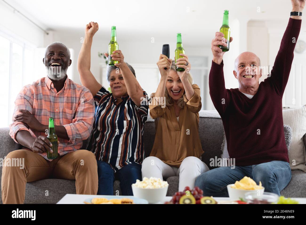 Coppie caucasiche e afroamericane senior che si siedono sul divano birra da gioco a casa Foto Stock