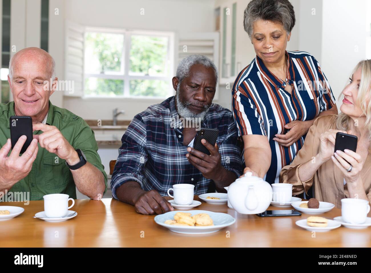 Coppie caucasiche e afroamericane senior seduti a tavola bevendo tè utilizzando lo smartphone a casa Foto Stock