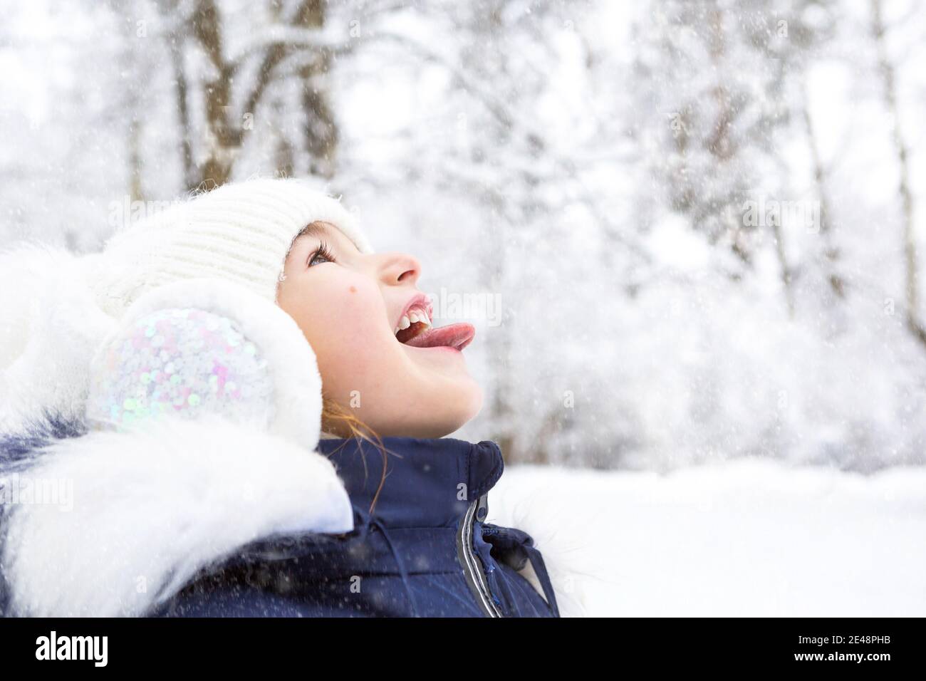 Una bambina con la lingua che pende fuori cattura fiocchi di neve in  inverno in una foresta nevosa. Cappello bianco lavorato a maglia, cuffie in  pelliccia con paillettes. Chi Foto stock -