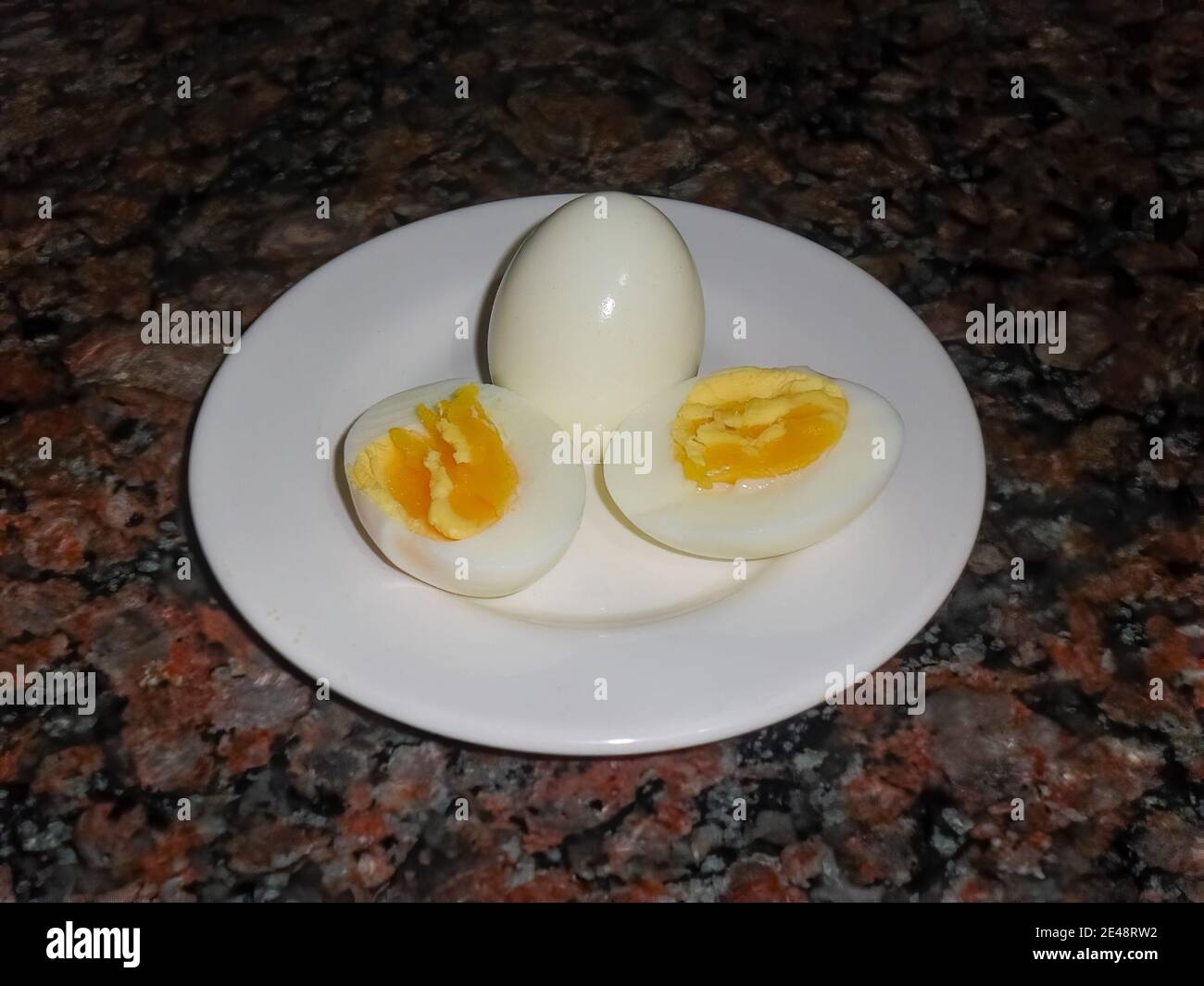 Due uova bollite su un piatto bianco, una a metà. Mostrando tuorlo. Sfondo di granito scuro. Angolo alto Foto Stock