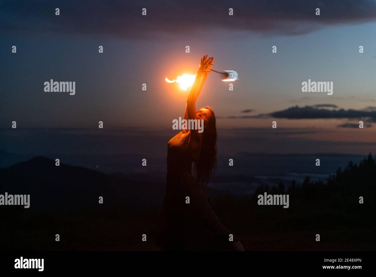 Aggraziata ed elegante artista femminile dei capelli rossi che fa la danza del fuoco con due ventilatori in metallo illuminati con fiamme sulle montagne notturne prato Foto Stock