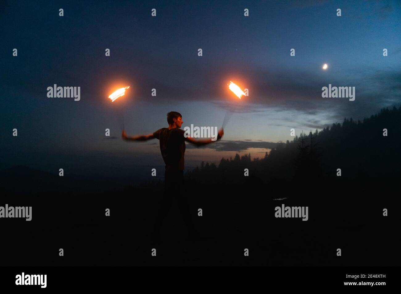Aggraziato performer di fuoco maschile che fa danza del fuoco con due metalli ventilatori illuminati con fiamme sul prato notturno delle montagne Foto Stock