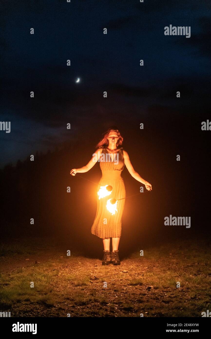 Aggraziata ed elegante artista femminile dei capelli rossi che fa la danza del fuoco con due ventilatori in metallo illuminati con fiamme sulle montagne notturne prato Foto Stock