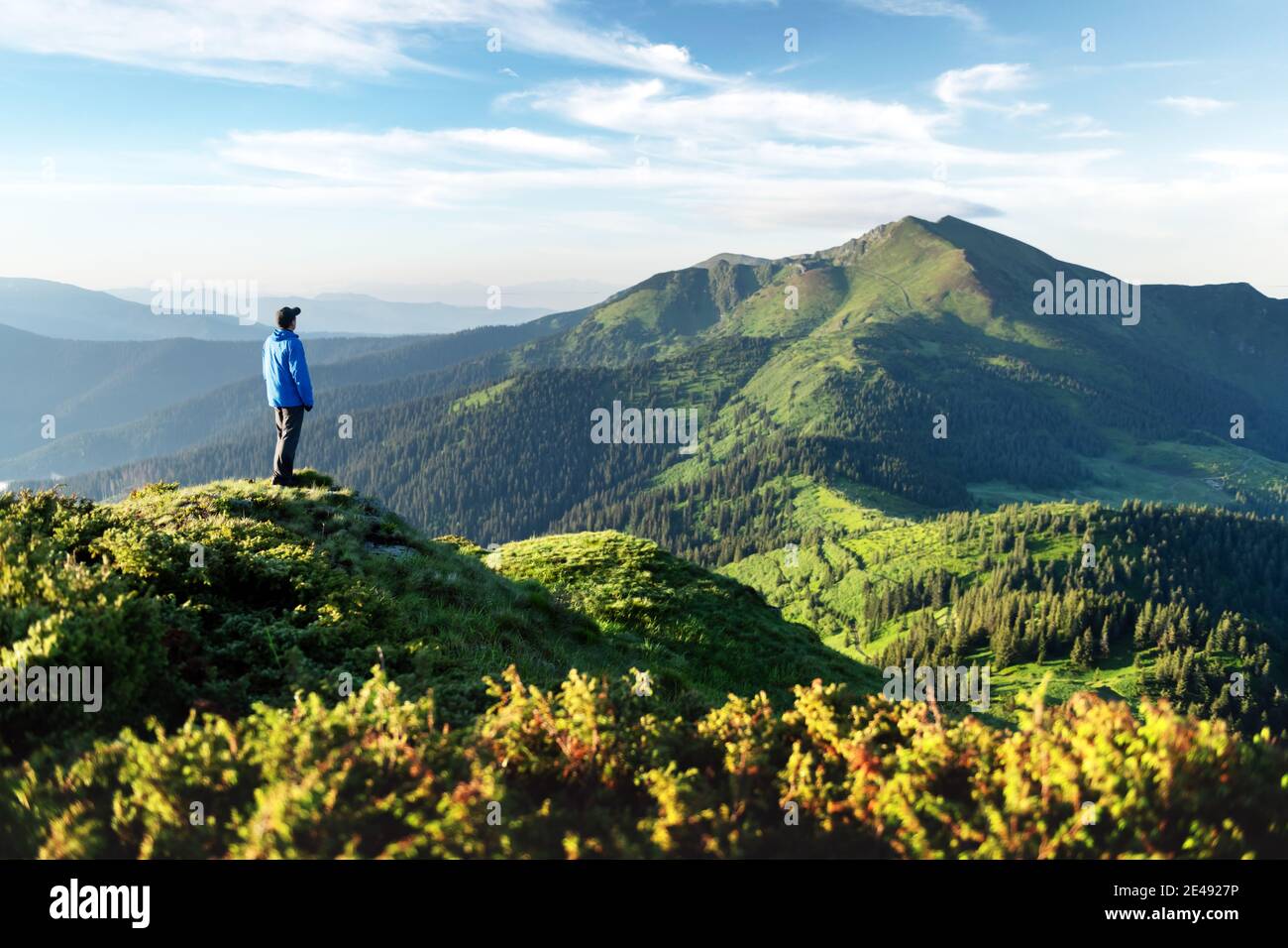 Un turista sul bordo di una montagna coperta da un'erba lussureggiante. Cielo blu nuvoloso e altissime montagne sullo sfondo. Fotografia di paesaggio Foto Stock