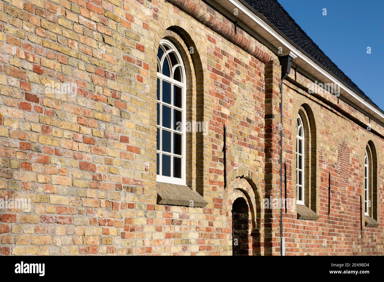 Facciata laterale di una vecchia chiesa restaurata con finestre ad arco e una porta in una muratura di vecchi mattoni gialli, arancioni e rossi contro un cielo blu in Exmorra Foto Stock