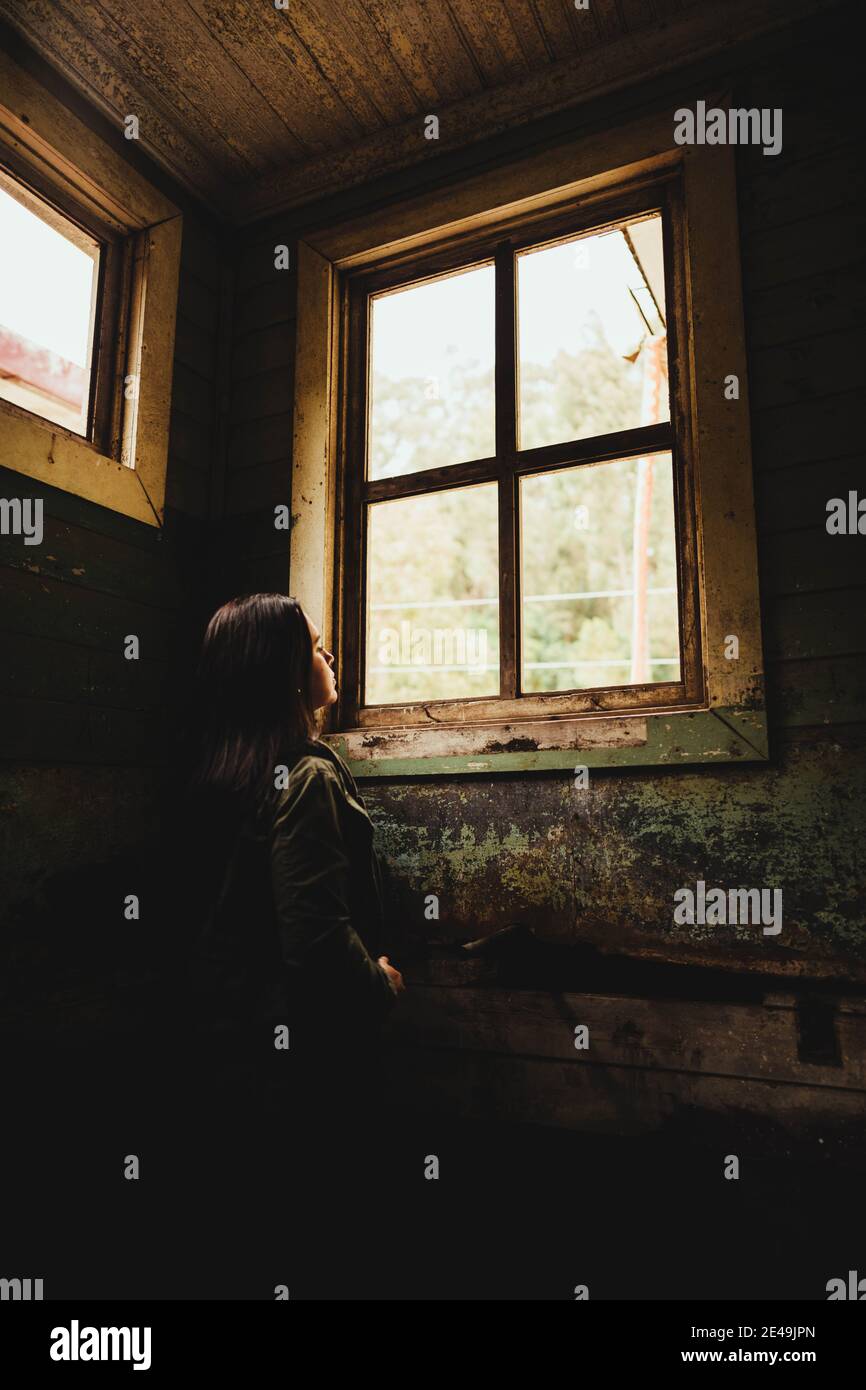 Donna che guarda attraverso la finestra indossando casual in un vecchio camera Foto Stock