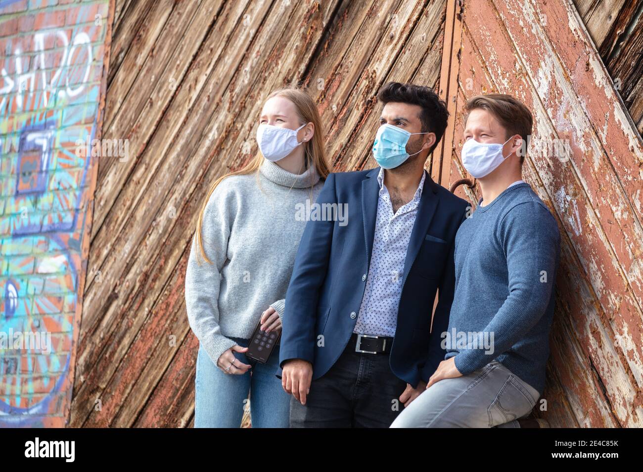 Gruppo di giovani, tempo libero in città. In piedi all'aperto. Maschera per bocca e naso attivata. Tre di loro si incontrano nello spazio pubblico. Requisito maschera. Foto Stock