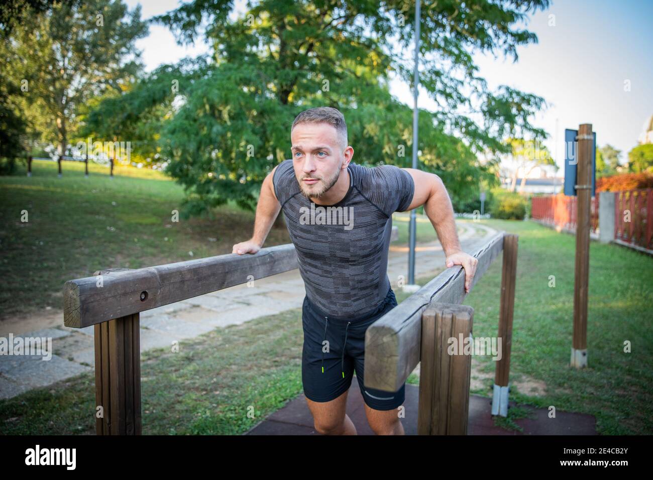 Uomo europeo in pantaloncini neri e t-shirt grigia che si tuffa su barre parallele. Allenamento all'aperto in un parco Foto Stock