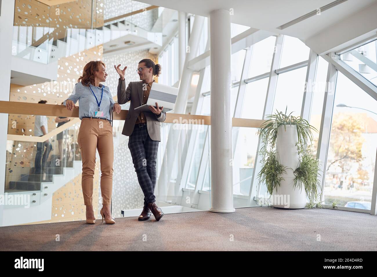 giovane che spiega alla collega anziana che si trova nel corridoio di un edificio commerciale, appoggiata su una recinzione, Foto Stock