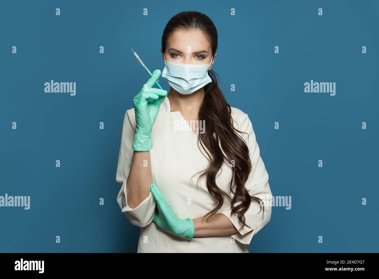 Medico o infermiere bella donna in maschera medica protettiva che tiene la siringa in piedi su sfondo blu. Concetto di medicina, sicurezza e vaccinazione Foto Stock
