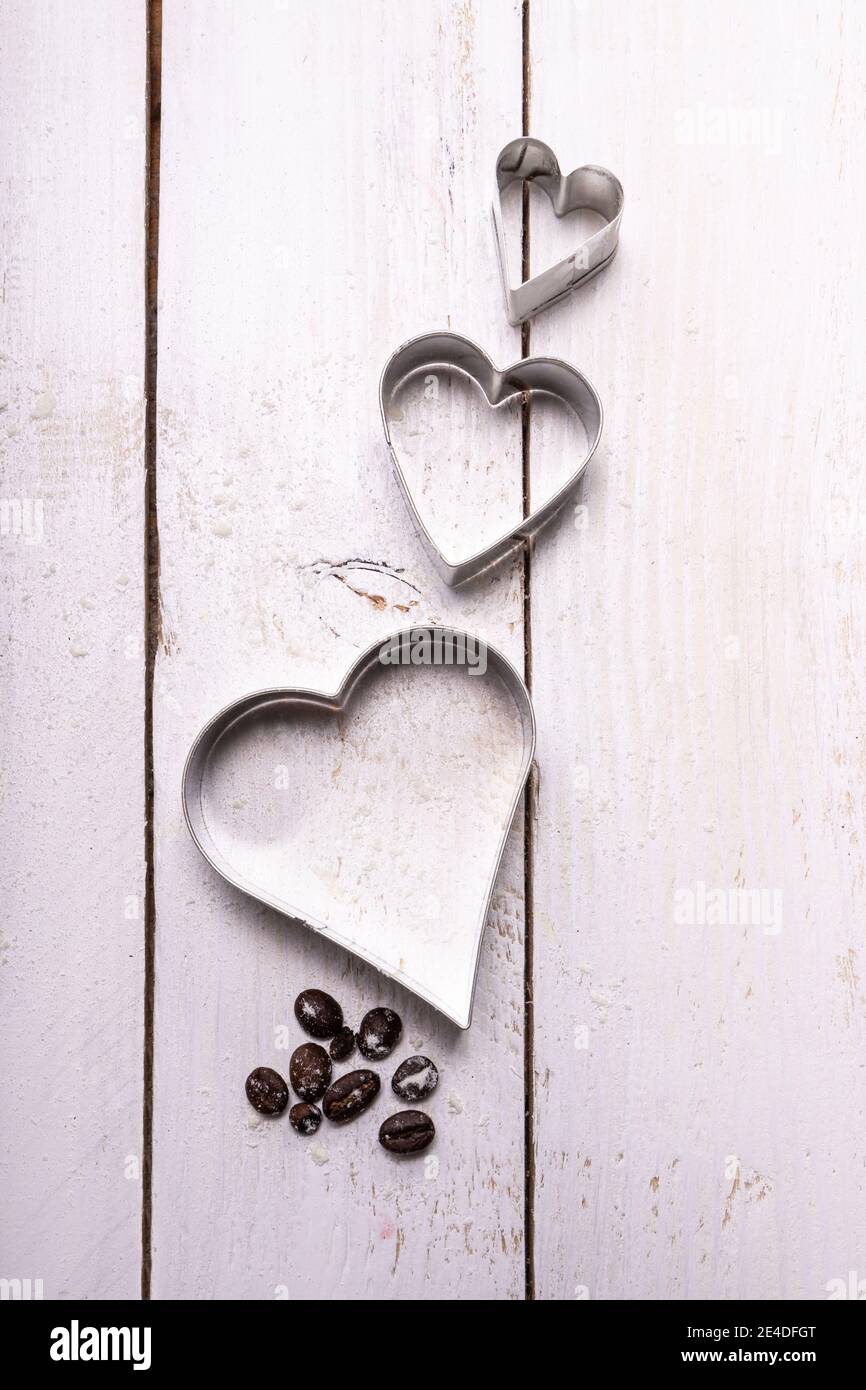 Composizione di stampi per biscotti a forma di cuore con alcuni chicchi di caffè su un pannello di legno bianco. Lascia che il tuo amore fliggh. Foto Stock
