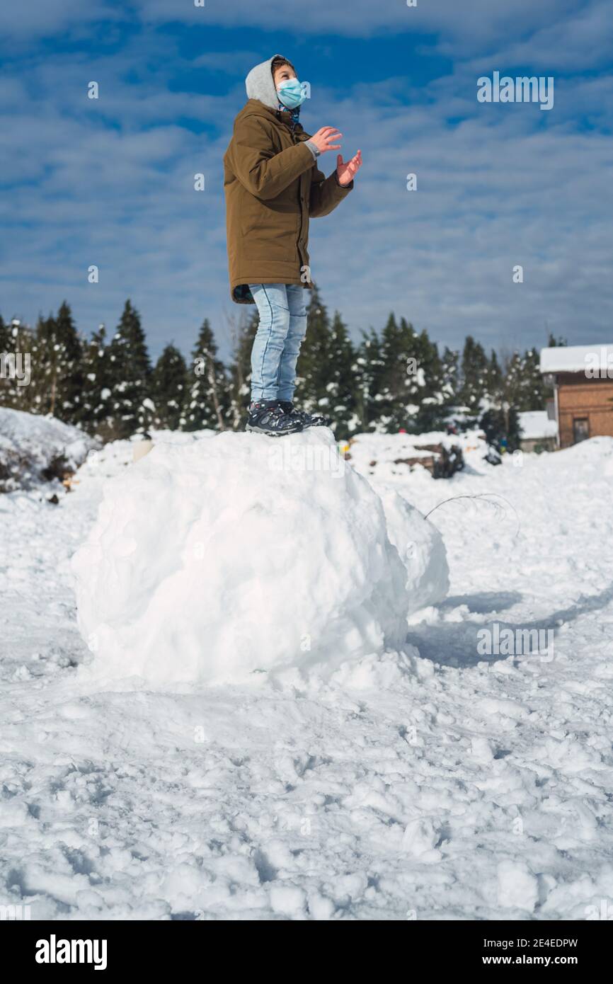 Caucasico ragazzo con una maschera in piedi su una grande palla di neve e giocare - vacanze invernali, pandemia Foto Stock