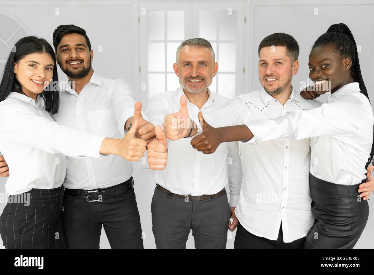 Gruppo di felici e sorridenti impiegati internazionali in camicie bianche mostrare i pollici verso l'alto e guardare la fotocamera Foto Stock