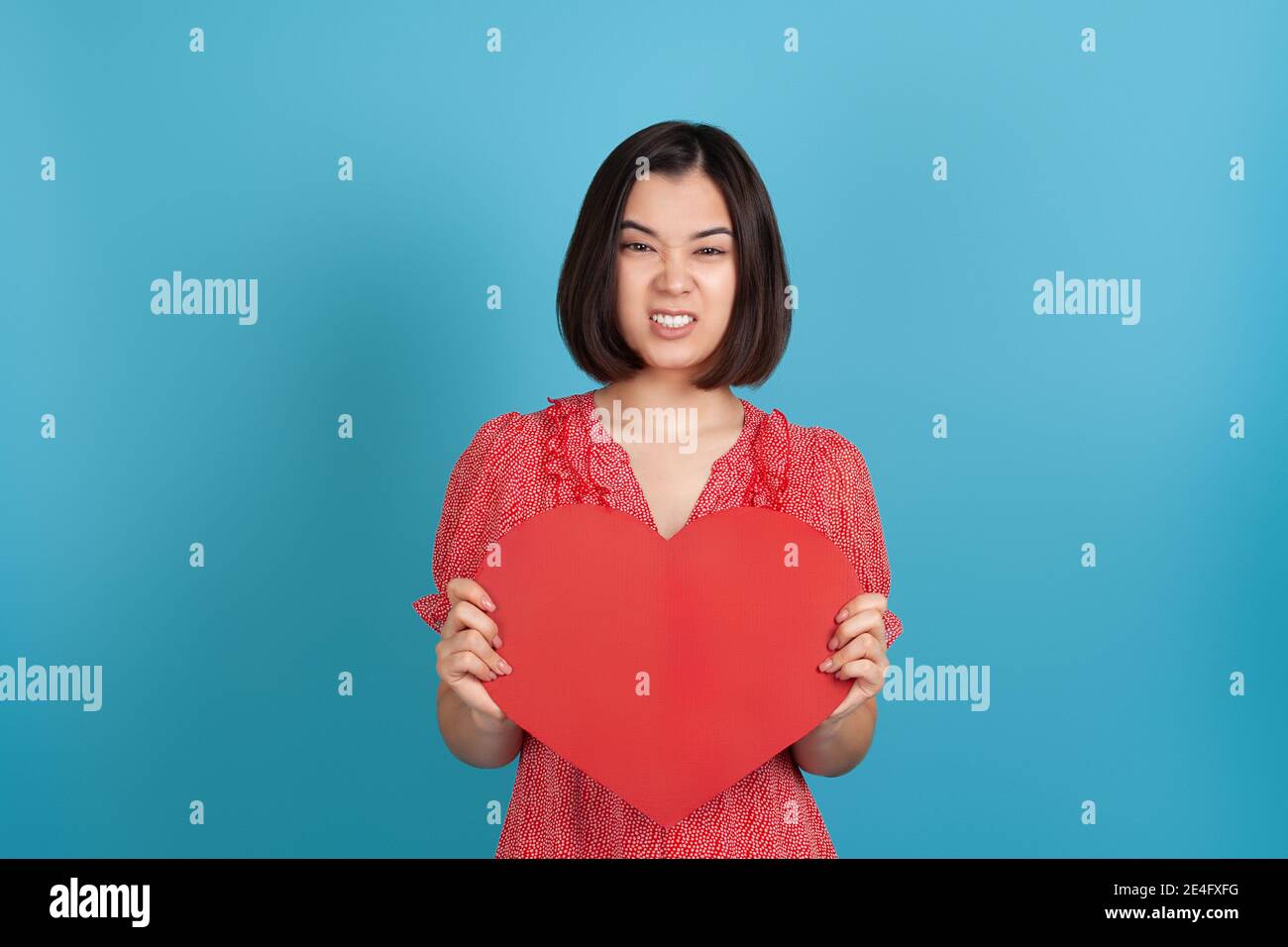 Primo piano offeso deluso giovane donna asiatica sconvolto in vestito rosso che tiene grande cuore di carta rossa e la sua denti, isolato su uno sfondo blu Foto Stock