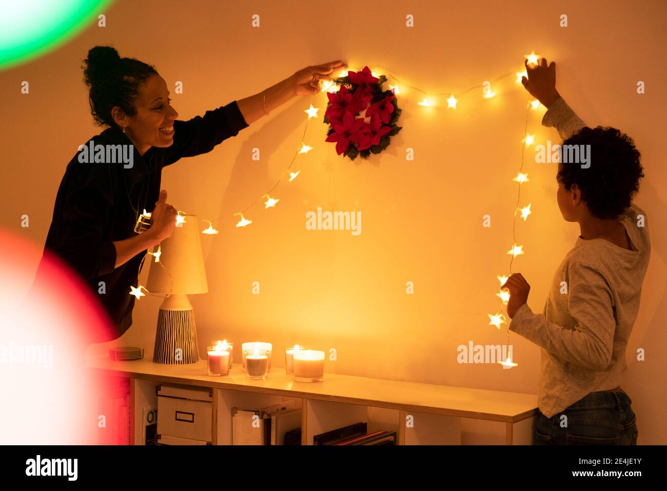 Madre sorridente con figlio che decorano luci di Natale illuminate sulla parete a casa Foto Stock