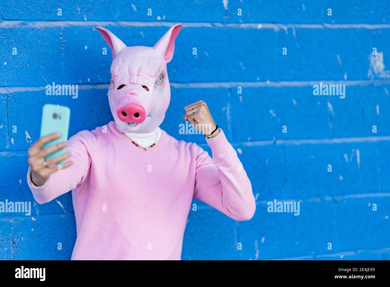 Giovane uomo in maschera di maiale mostrando pugno mentre prendendo selfie contro parete blu Foto Stock