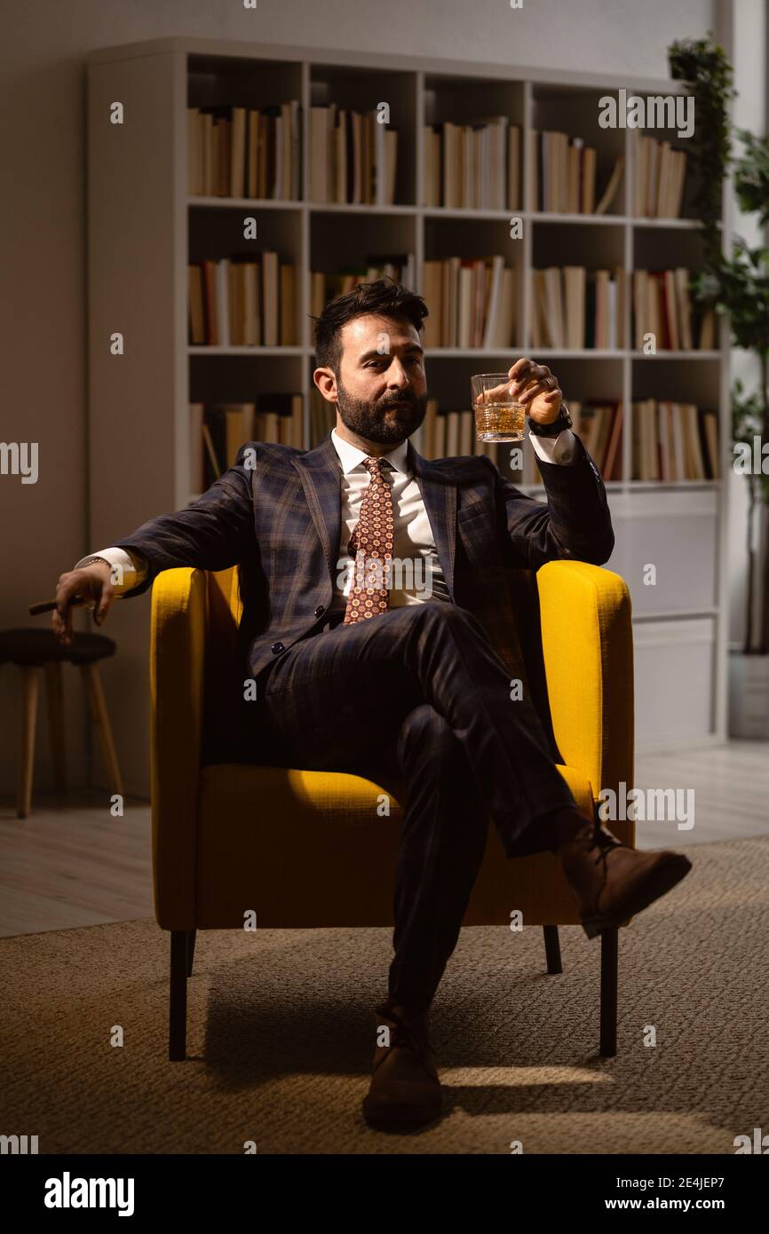 Ritratto di uomo ben vestito seduto in poltrona e godendo di sigaro e un bicchiere di whisky Foto Stock
