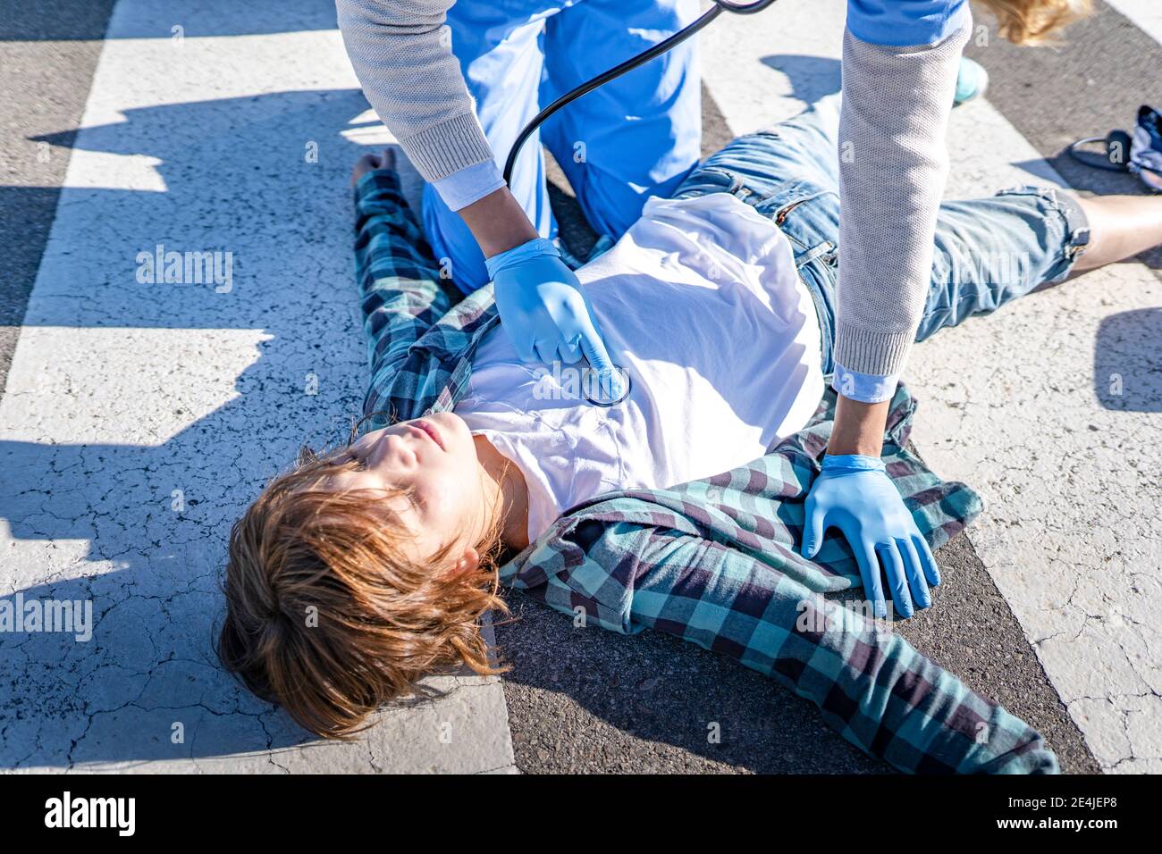 Operaio di salvataggio maschile con stetoscopio che esamina ragazzo sdraiato su strada dopo un incidente stradale Foto Stock