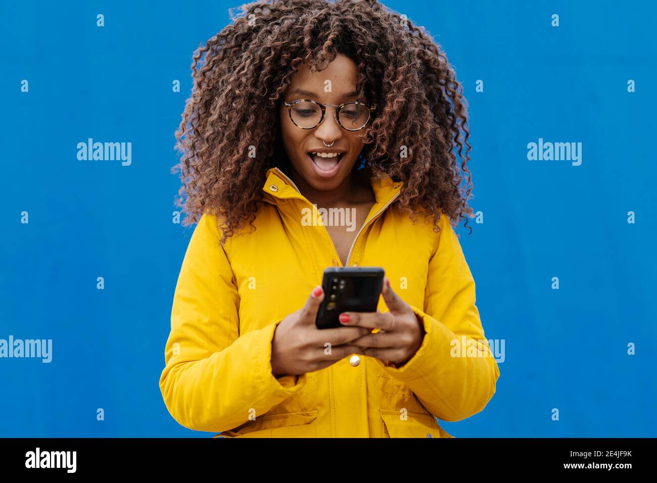 Donna sorpresa con bocca aperta utilizzando il telefono cellulare mentre si è in piedi contro parete blu Foto Stock