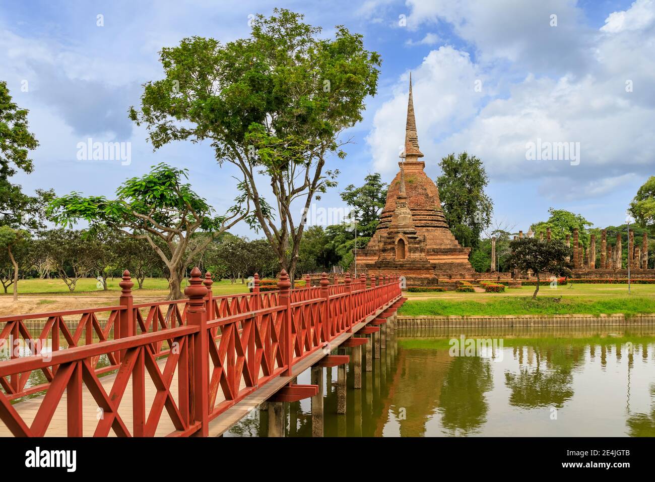 Ponte di legno rosso attraverso lo stagno che conduce alla pagoda e rovinato complesso monastery al tempio di Wat SA si, Sukhothai Historical Park, Thailandia Foto Stock