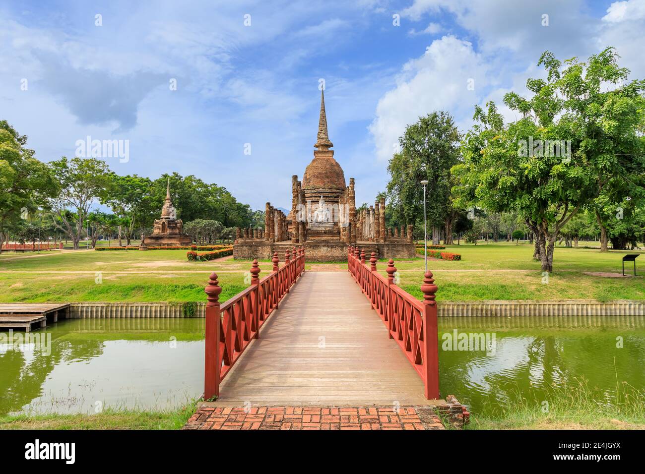 Ponte di legno rosso attraverso lo stagno che conduce alla pagoda e al complesso di cappella in rovina monastero al tempio di Wat SA si, Sukhothai Historical Park, Thailandia Foto Stock