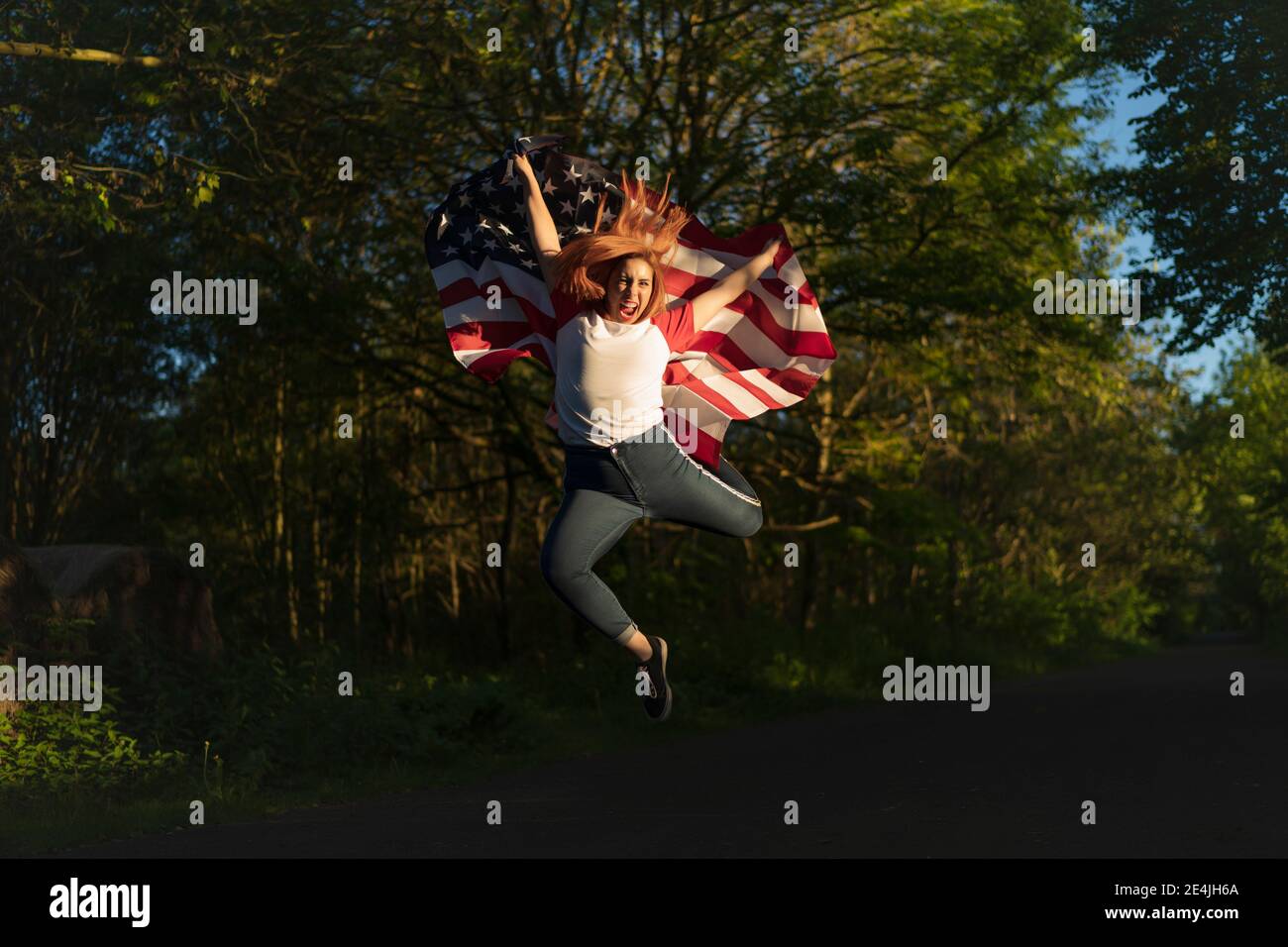 Donna spensierata che tiene la bandiera americana mentre saltando sulla strada durante tramonto Foto Stock