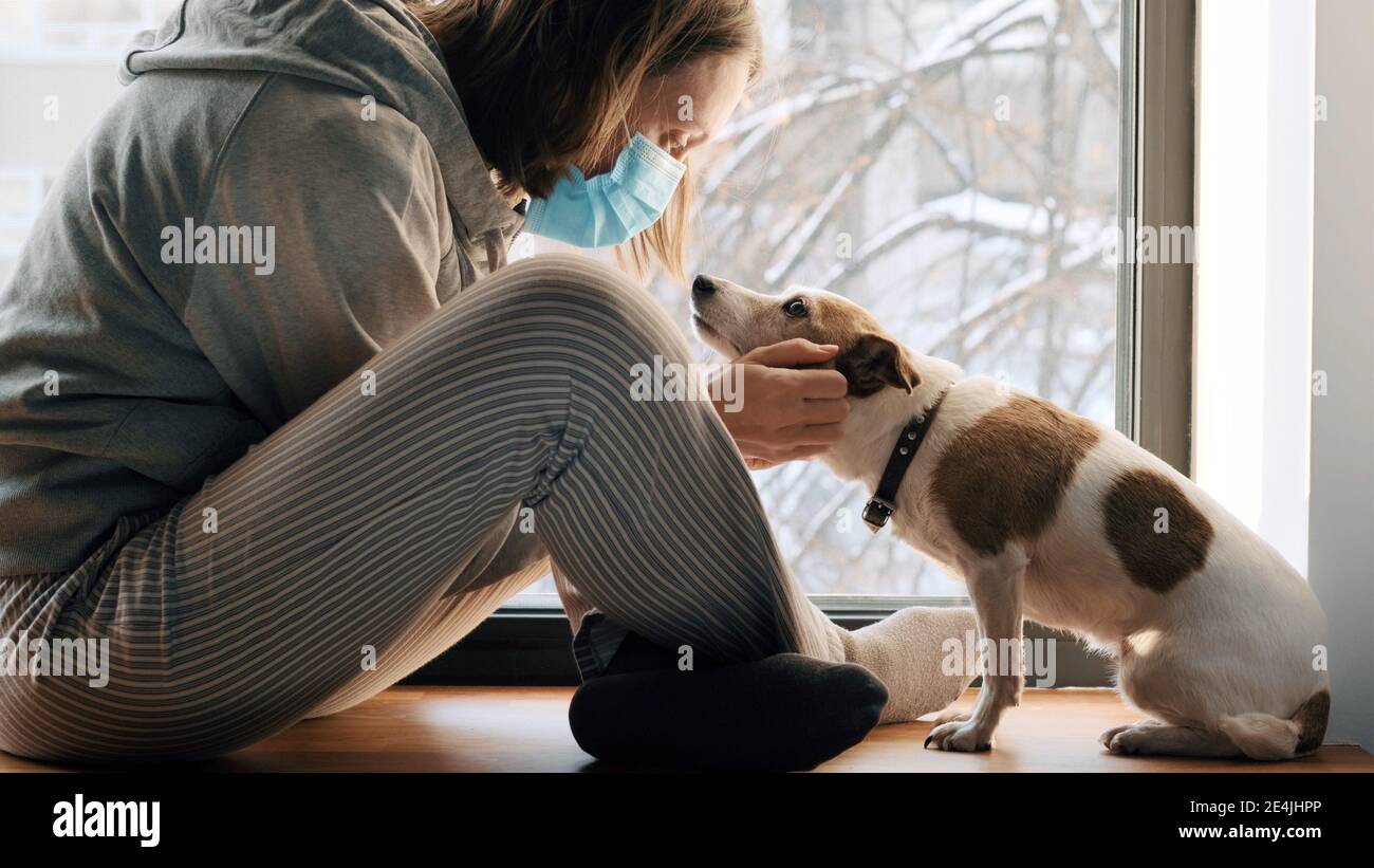 Donna seduta in maschera medica alla finestra con un cane shaggy. Concetto di autoisolamento virus covid 19 infezione doomsda coronavirus Foto Stock