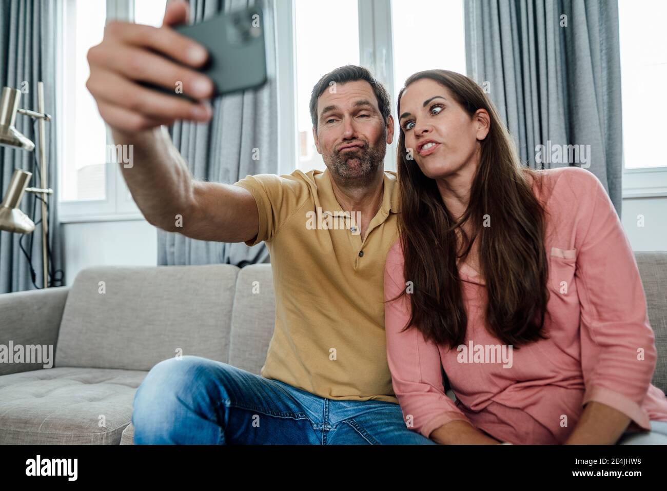 Coppia felice che fa i volti mentre prende selfie mentre si siede sopra divano a casa Foto Stock